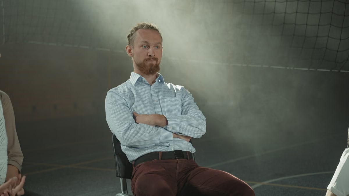 A man sits pensively during a group therapy session, illuminated by soft, filtered light indoors.